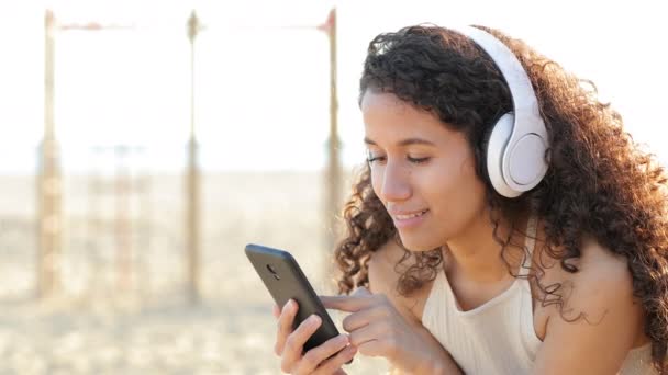 ラテン系の女性はビーチで屋外のスマートフォンから音楽を聞いて感じます — ストック動画