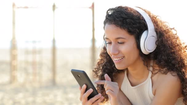 音楽を聞いてヘッドフォンを身に着けているラテン女性スマートフォンをチェック上の座っ — ストック動画