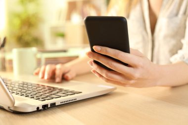 Kadın eli kapalı, akıllı telefonu kontrol ediyor evdeki bir masada dizüstü bilgisayar kullanıyor. 