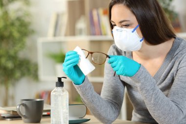Woman with protective mask and gloves disinfecting eyeglasses due coronavirus sitting at home clipart