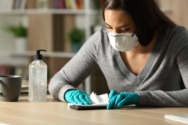 Eldivenli ve koruyucu maskeli bir kadın Coronavirus 'tan akıllı telefonu dezenfekte ediyor. Geceleri evdeki masada oturuyor.