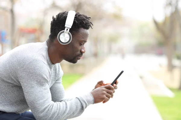 Profile of a serious black man with headphones listening music on smart phone sitting in a park bench