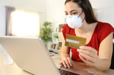 İnternette internetten ödeme yapan kadın bilgisayarında maske takıyor. Evde bir masada oturuyor.