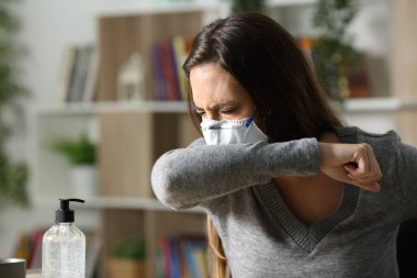 Maskeli hasta kadın, gece evde dirseğinde Coronavirus enfeksiyonu yüzünden öksürüyor.