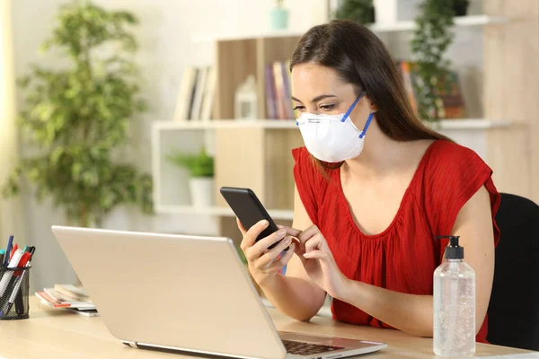 Woman checking smart phone avoiding coronavirus with mask sitting on a desk at home