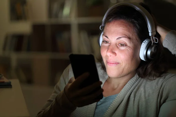 ヘッドフォンを持つ幸せな大人の女性は自宅で夜に座って点灯画面とスマートフォンで音楽を聴く — ストック写真