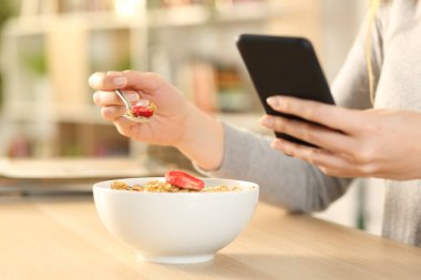 Kadın ellerinin mısır gevreği kasesi ve meyve yerken evdeki masada oturan akıllı telefonları kontrol etmesi.
