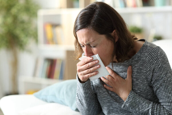 Больная взрослая женщина кашляет, закрывая рот салфеткой, сидя дома на диване
