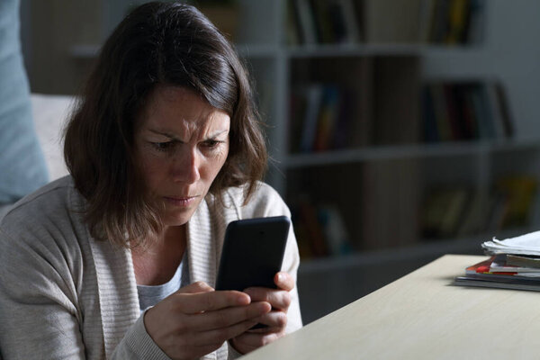 Worried adult woman reading bad news on smart phone sitting on the floor at night at home