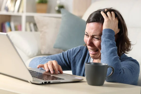 Sad adult woman crying reading bad news on laptop sitting on the floor on a coffee table at home