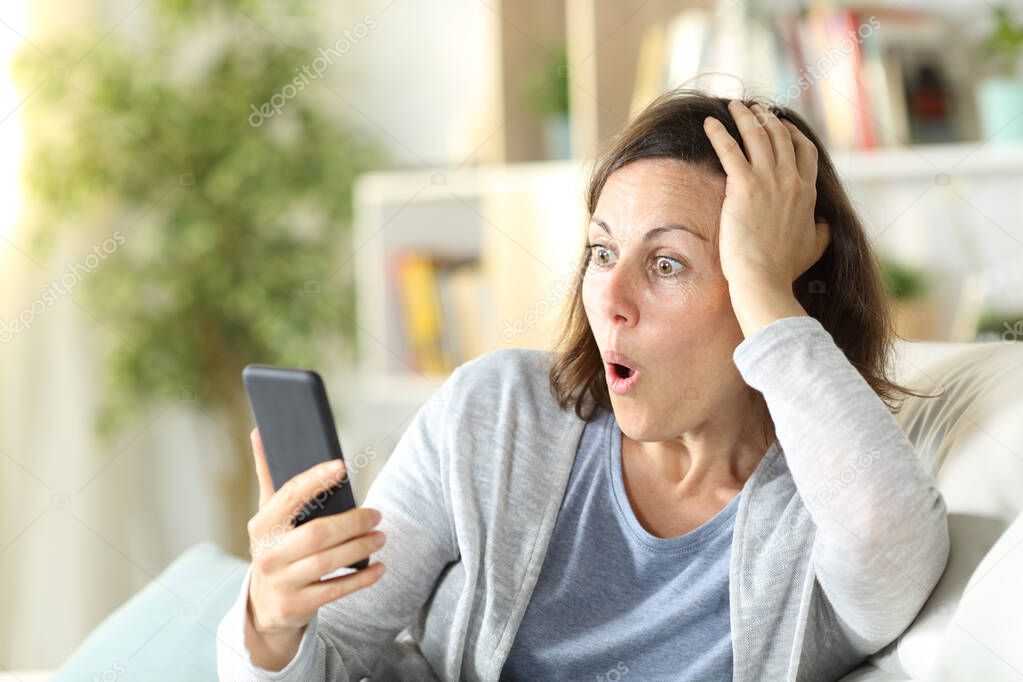 Surprised adult woman reading news on smart phone sittng on a couch at home
