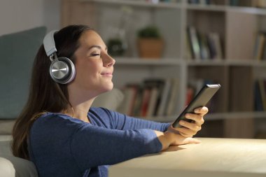 Mutlu kız gece evde oturan kulaklıkla akıllı telefondan müzik dinliyor.