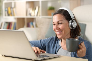 Happy adult woman with headphones watching video online on laptop sitting on the floor at home holding coffee cup clipart