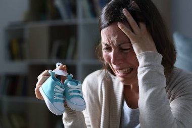 Sad mother crying missing her daughter after miscarriage holding new baby shoes at night at home clipart