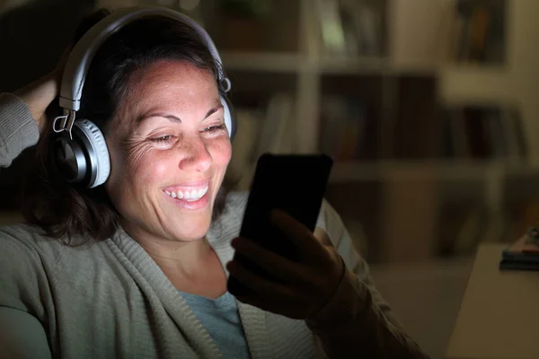 幸せな大人の女性とヘッドフォンは自宅で夜のリビングルームに照明付きスマートフォンで音楽を聴く — ストック写真