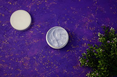 Beyaz kapta yüz ya da vücut kremi. Organik doğal bitkisel kozmetik, cilt için nemlendirici. Mor arka planda çiçekler. Üst görünüm, alanı kopyala.