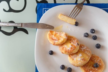 Syrniki ya da yaban mersinli süzme peynir krepleri ve çatal ve bıçakla mutfak masasında mavi peçete ile beyaz tabakta bal. Sağlıklı kahvaltı, diyet konsepti.