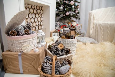 Noel salonunun içi süslenmiş köknar ağacı, şömine, beyaz koltuk, tüylü halı, hediyeler, kozalaklı sepetler.