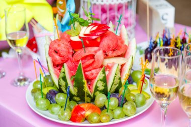 Meyve tabağı, karpuz oyması ve ön planda odaklanmamış şampanya bardakları olan şenlikli çikolata..