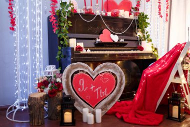Sevgililer Günü konsepti. Piyano, mumlar, kırmızı tahta kalpler ve çiçeklerle süslenmiş. Kırmızı ekose ve yazılı odaksız sandalye Ben + Sen ön planda.