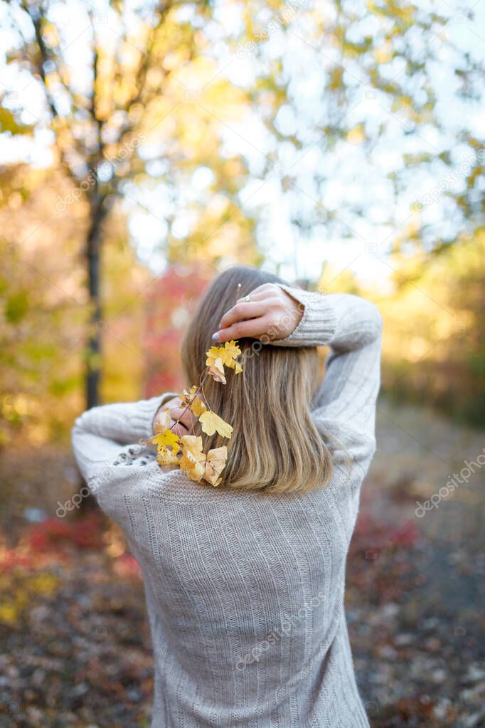 girl in knit sweater standing with her back to camera holding a branch with leaves near hair in the autumn forest