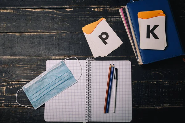 Notebook, alphabet cards, pencils and medical gauze mask on wooden table. Covid 2019, selfisolation, distant learning, home education, remote study concept. Copy space.