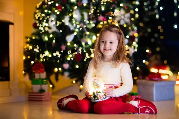 Kar küresi Noel ağacının altında tutan küçük kız — Stok fotoğraf