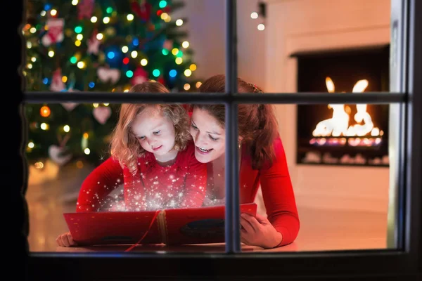 Mother and daughter read a book at fireplace on Christmas eve.