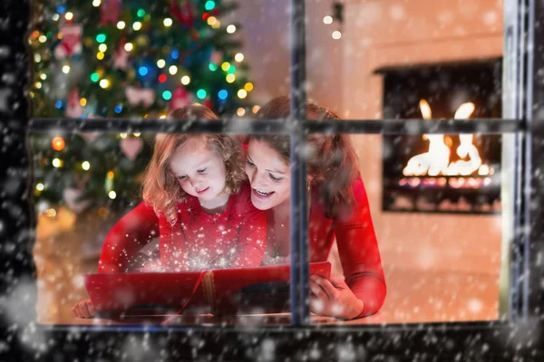 Mother and daughter read a book at fireplace on Christmas eve.