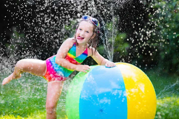 Rolig skrattande liten flicka i en färgsprakande badkläder leker med leksak boll trädgård sprinkler med vatten splashe — Stockfoto