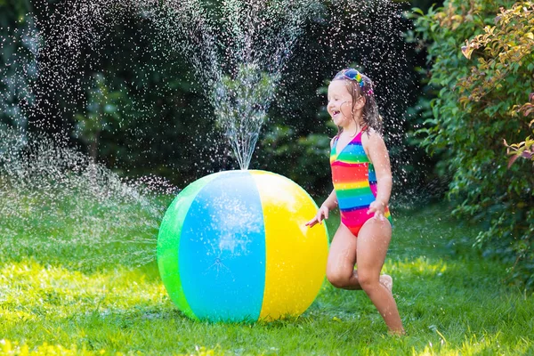 Komik küçük kız oyuncak top Bahçe yağmurlama ile oynarken gülüyor — Stok fotoğraf