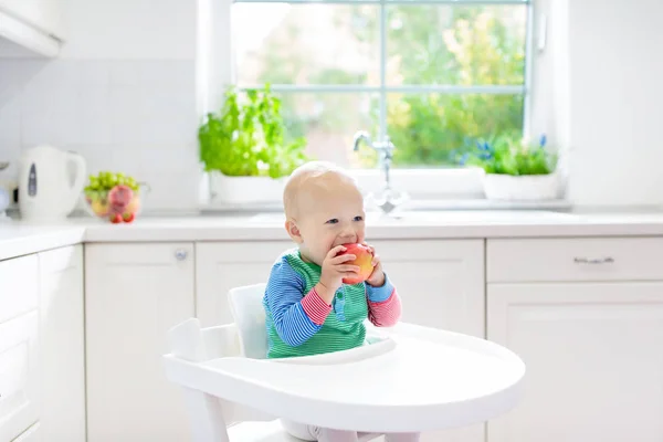 Baby boy eating apple in white kitchen at home — Stock Photo, Image