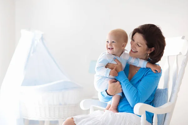 年轻的母亲抱着她刚出生的孩子 妈妈护理宝宝 女人和新出生的男孩 在白色卧室 摇椅和蓝色婴儿床 幼儿园的内部 母亲笑着孩子玩 家人在家里 — 图库照片