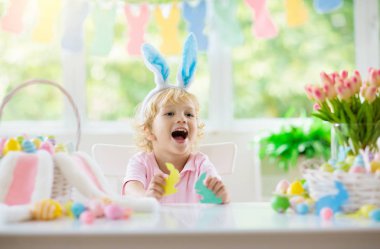 Çocuklar paskalya yumurtalarını boyuyor. Tavşan kulaklı çocuklar Paskalya avı için renkli yumurta boyuyorlar. Bahar tatili kutlaması için çiçek, sepet ve tavşanlı ev dekorasyonu. Küçük bebek evi dekore ediyor..