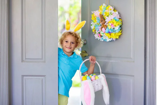 Kids at Easter egg hunt. Child holding basket with colorful chocolate eggs, wearing bunny ears. Little boy at decorated front door after Easter celebration. Children celebrate spring holiday.