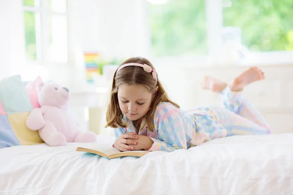 窓付きの白い日当たりの良いベッドルームでベッドで子供の読書本 子供は本を読む キッズルームとインテリアデザイン パジャマ姿の女の子が家にいる 保育園のための寝具と寝具 おもちゃ付きの子供 — ストック写真