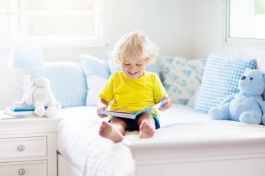 Beyaz güneşli bir odada pencereli bir çocuk yatakta oynuyor. Çocuk odası ve iç tasarım. Erkek bebek evde. Çocuk yuvası için yatak ve tekstil. Oyuncağı ve kitabı olan bir çocuk. Uyku ve uyku zamanı..