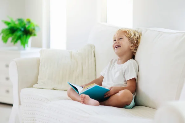 儿童读物 孩子们读书 小男孩坐在阳光明媚的客厅里的白色沙发上 看着故事书里的照片 孩子为小学或幼儿园做作业 儿童学习 — 图库照片