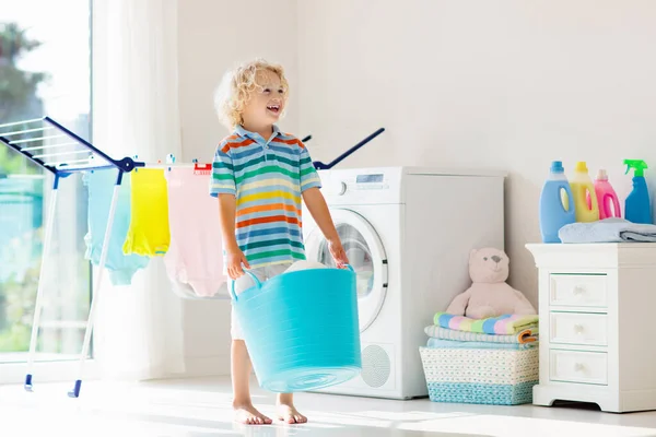 Child in laundry room with washing machine or tumble dryer. Kid helping with family chores. Modern household devices and washing detergent in white sunny home. Clean washed clothes on drying rack.