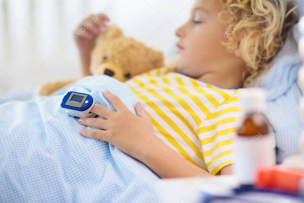 Sick little boy with pulse oximeter on his finger. Asthma treatment. Ill child lying in bed. Unwell kid with chamber inhaler, cough medicine. Flu season. Bedroom or hospital room for young patient. 