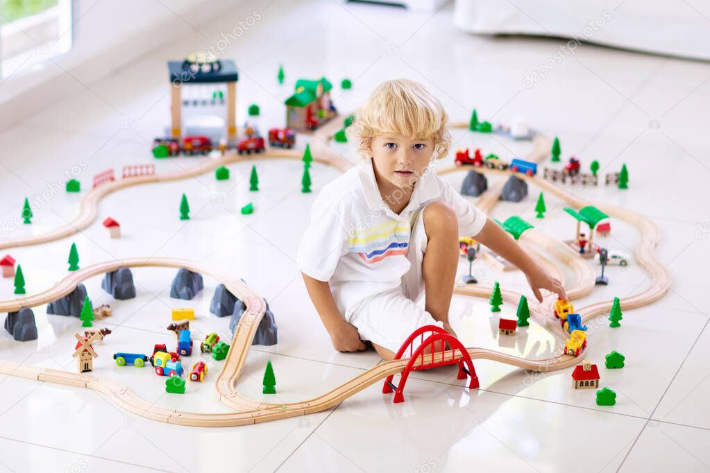 Kids play with wooden railway. Child with toy train. Educational toys for young children. Little boy building railroad tracks on white floor at home or kindergarten. Cute kid playing cars and engine.