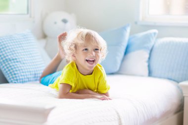 Beyaz güneşli bir odada pencereli bir çocuk yatakta oynuyor. Çocuk odası ve iç tasarım. Erkek bebek evde. Çocuk yuvası için yatak ve tekstil. Oyuncağı ve kitabı olan bir çocuk. Uyku ve uyku zamanı..