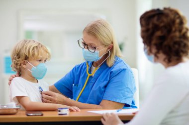 Çocuk doktoru, yüz maskeli hasta çocuğu muayene ediyor. Test ve tarama için sağlık kliniğindeki hasta çocuk. Çocuklar evde virüs tedavisi görüyor. Koronavirüs salgını. Covid-19 salgını. Hasta öksürüyor..