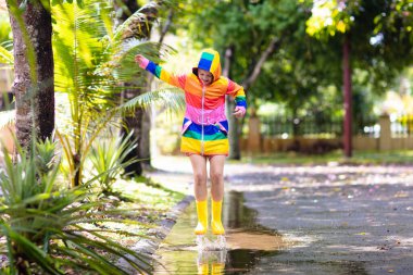 Sonbahar parkında yağmurda oynayan bir çocuk. Yağmurlu bir sonbahar gününde çamurlu su birikintisine atlayan bir çocuk. Yağmurluk ve gökkuşağı ceketli küçük kız dışarıda, sağanakta. Çocuklar su geçirmez ayakkabı ve ceket giyiyor..