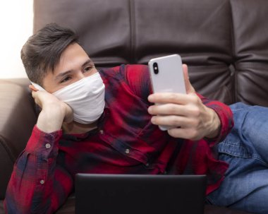 Koruyucu maskeli genç adam akıllı telefonuyla içeride karantina için etkileşime geçiyor. Covid-19 için kendini yalıtım