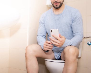 Yetişkin bir adam evde tuvalette otururken akıllı telefon kullanıyor. Teknoloji ve yaşam tarzı kavramı.