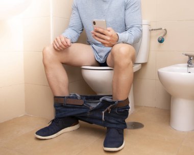 Yetişkin bir adam evde tuvalette otururken akıllı telefon kullanıyor. Teknoloji ve yaşam tarzı kavramı.