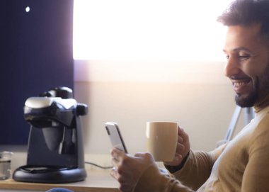 Genç bir adam kahve içiyor ve akıllı telefonundan mesaj okuyor.