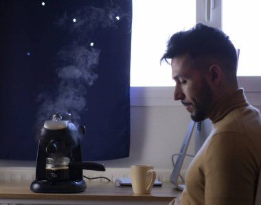 Yakışıklı bir genç adam kahve içiyor ve akıllı telefonuyla evde mesaj okuyor.