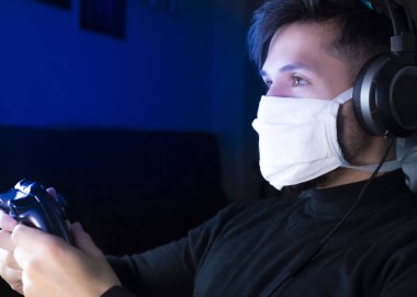 Covid-19 karantinaya alınırken genç beyaz adam yüzünde tıbbi maskeyle video oyunları oynuyor.
